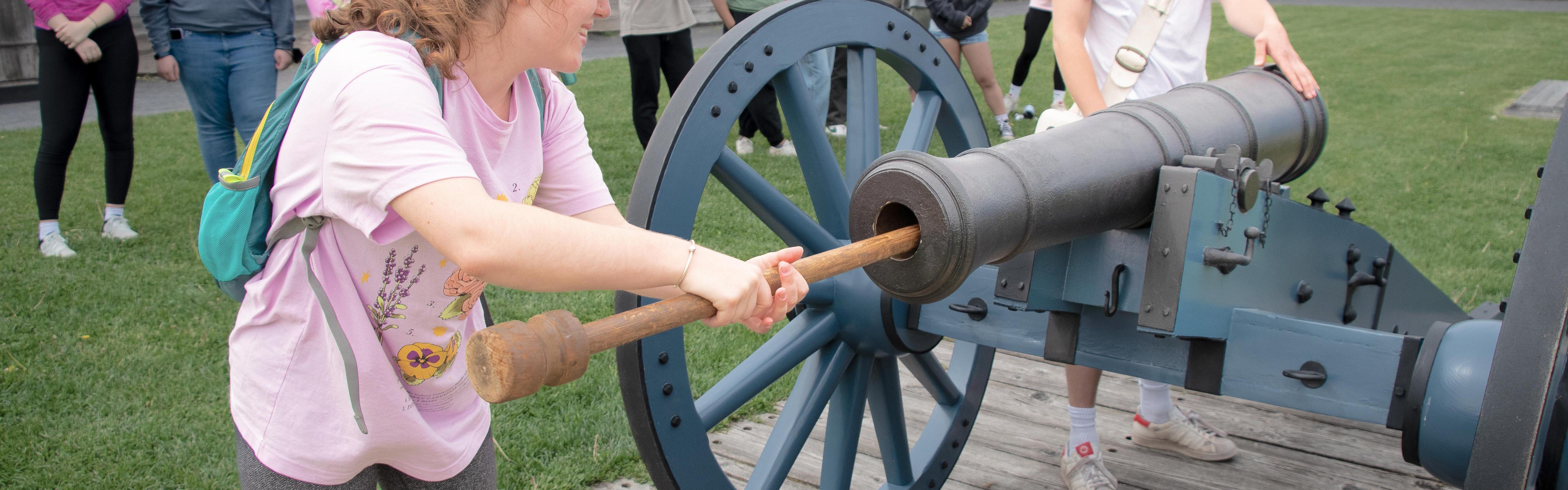 A young woman is using a long wooden q-tip like stick to clean a cannon while her friends watch on.