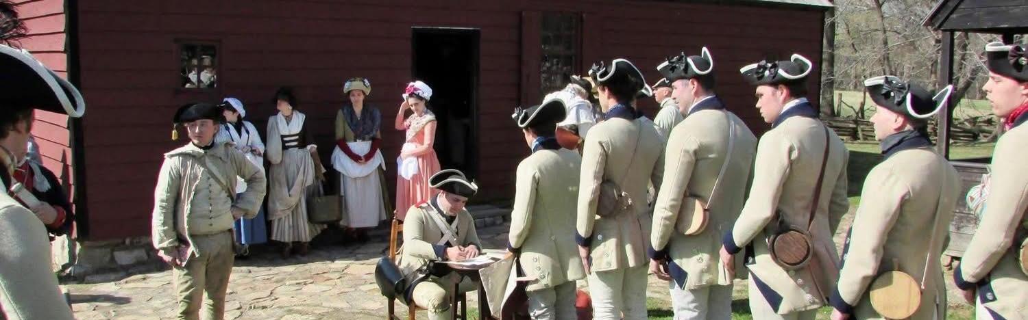 Men in grey Continental soldier uniforms stand in line near a large wooden building.