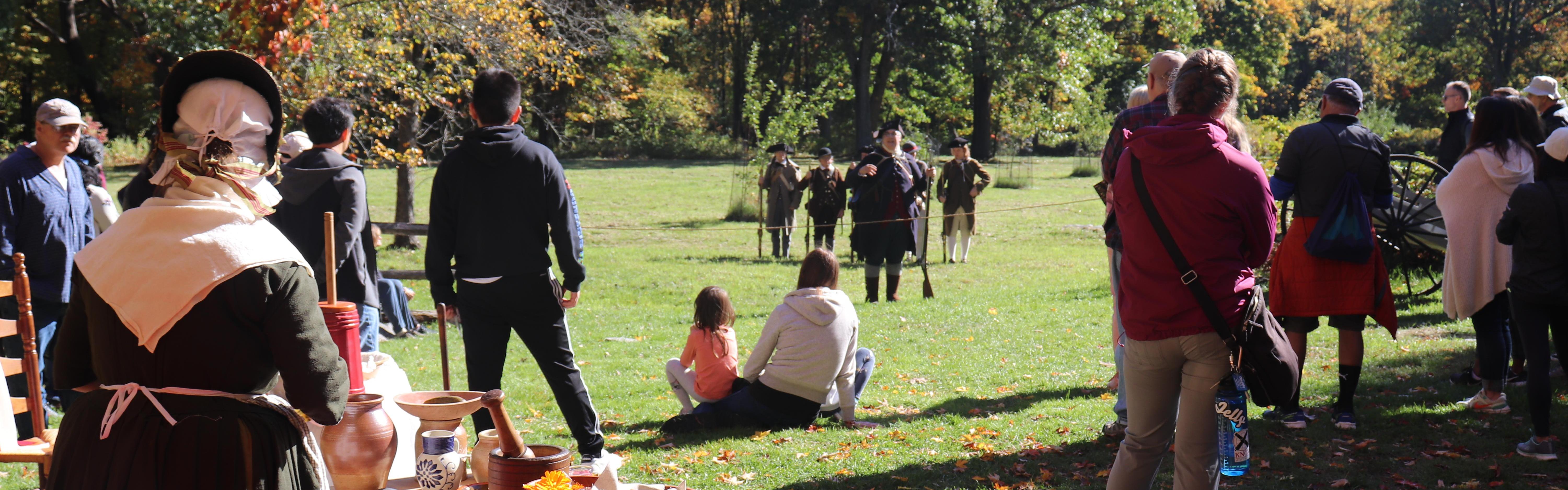 People in colonial clothing mingle with modern visitors in a field with trees in autumn color