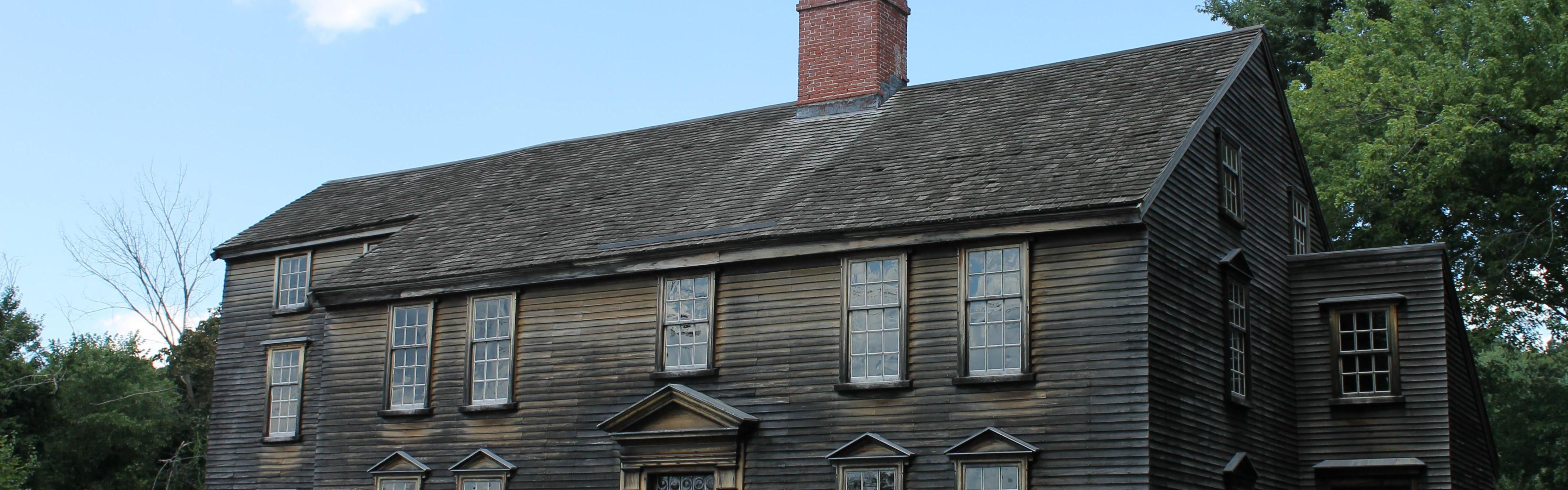 A three story colonial saltbox style house made of wood. It is surrounded by green grass and trees.