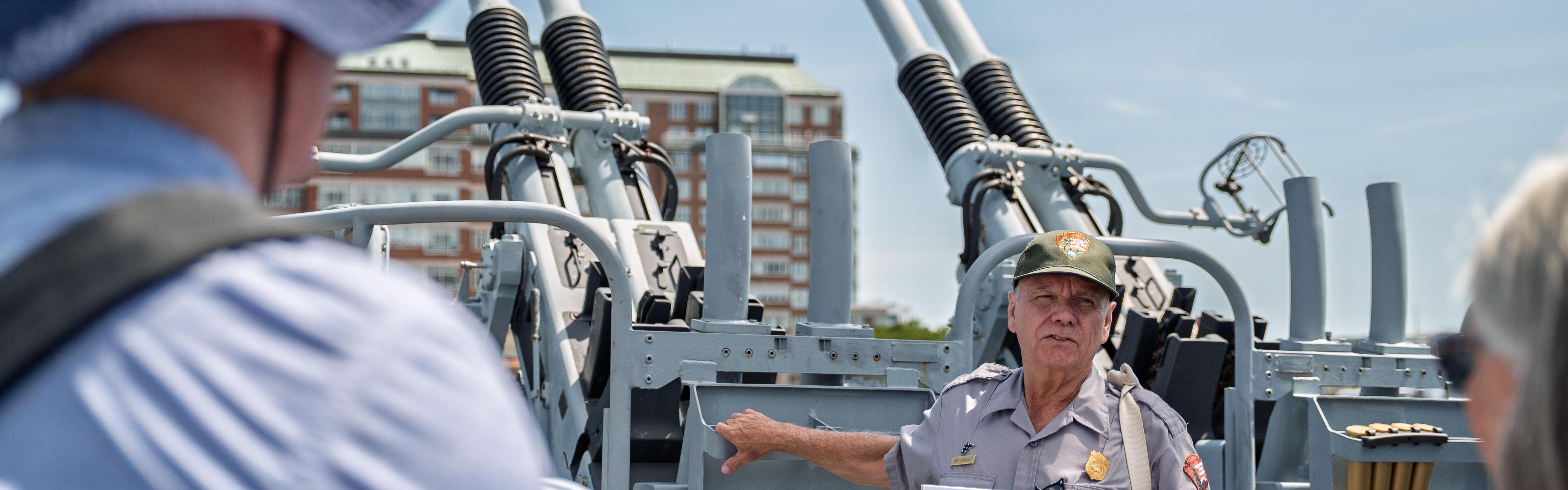 A ranger stands in front of a gun mount speaking to members of the public.