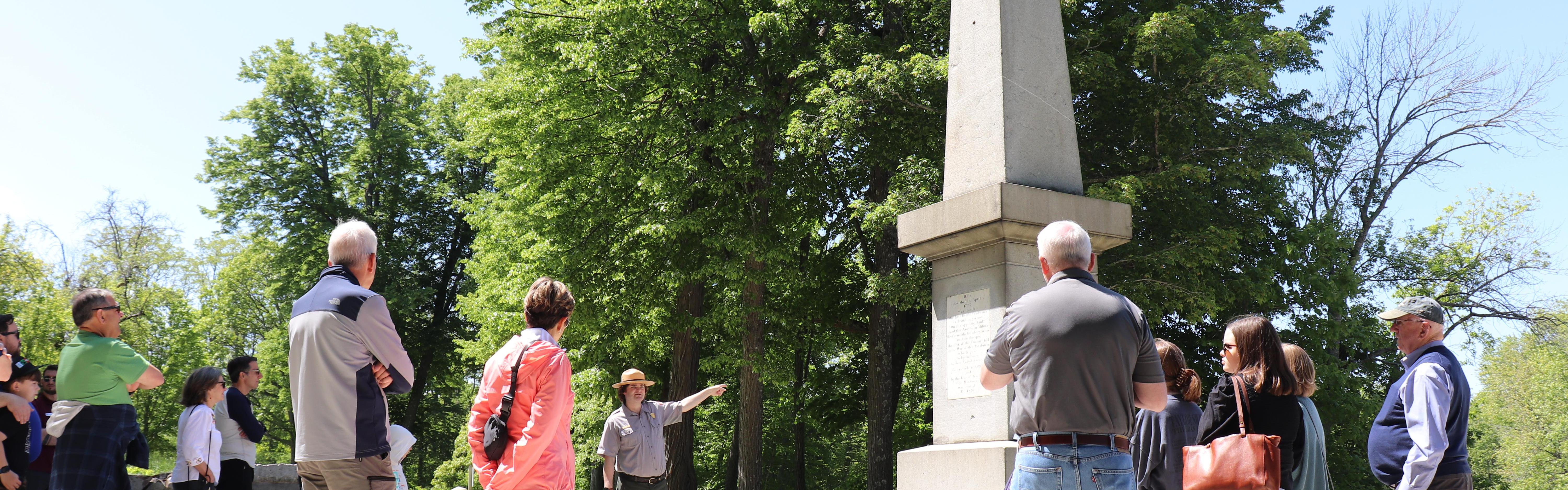 A park ranger stands in front of a towering stone monument pointing as visitors stand around.