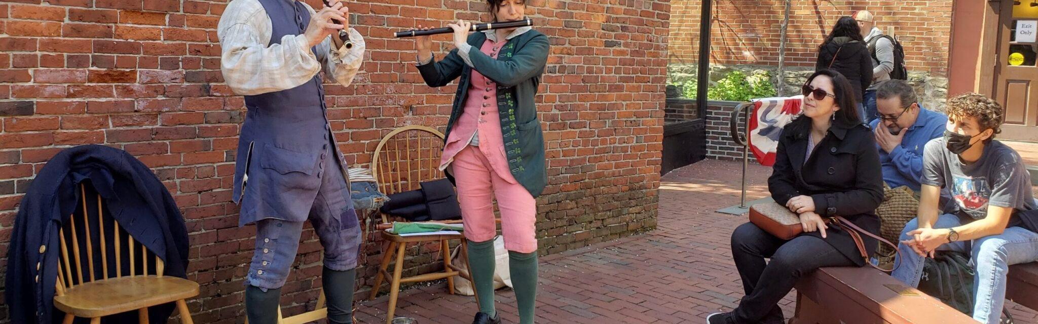 Two people wearing colonial garb and playing instruments while a few people watch