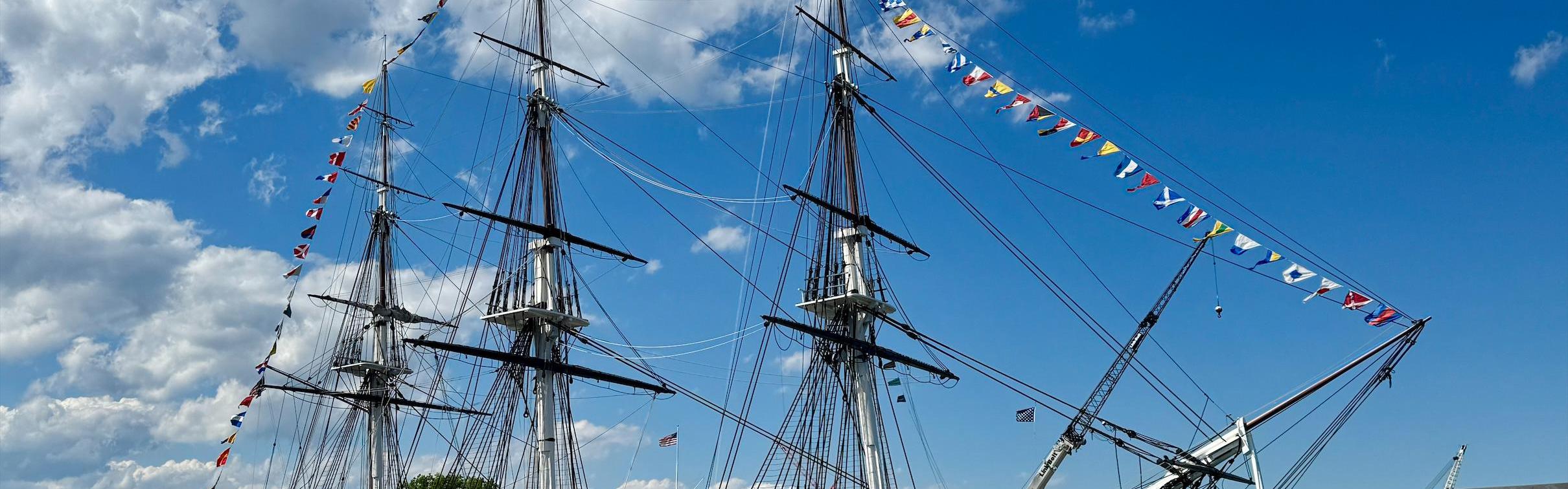 USS Constitution, a three-masted sailing ship, is decorated in flags on a partly-cloudy day.