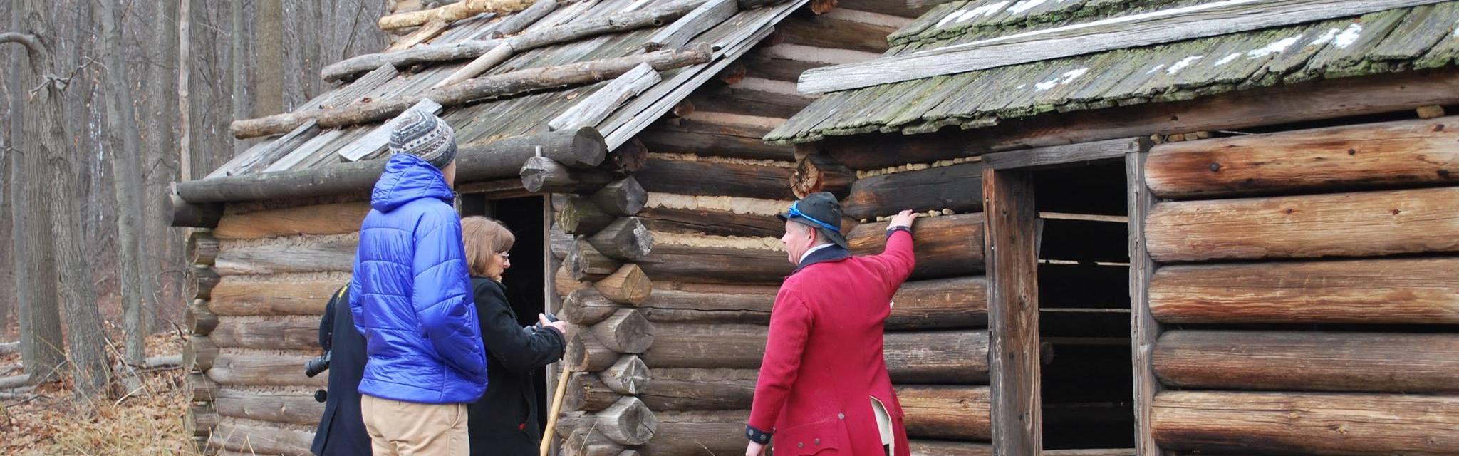 Two men in colonial living history clothing standing in front of log cabins.