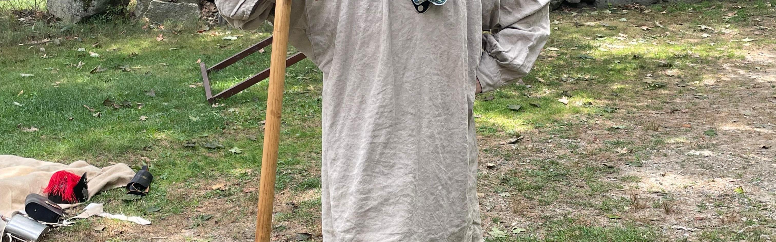 A colonial man with a cloth over his face stands holding a shovel