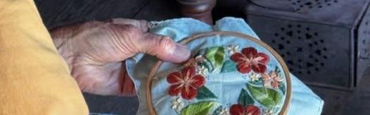 A volunteer in colonial dress practices cross stich. She is stitching a flower design.