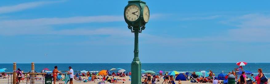 Green clock on boardwalk of ocean beach park site with people, umbrellas and blankets on sand