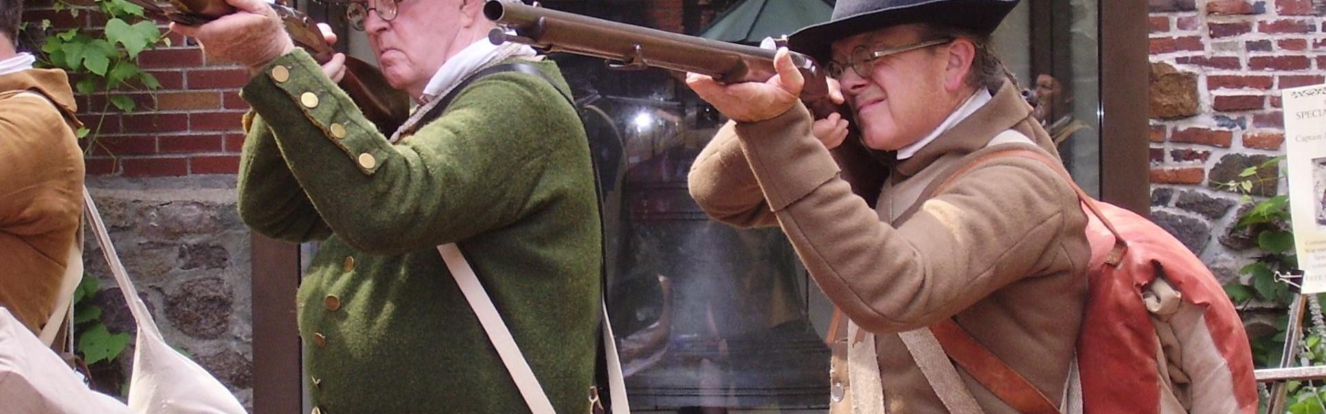 two men in colonial militia clothing pointing muskets at the camera