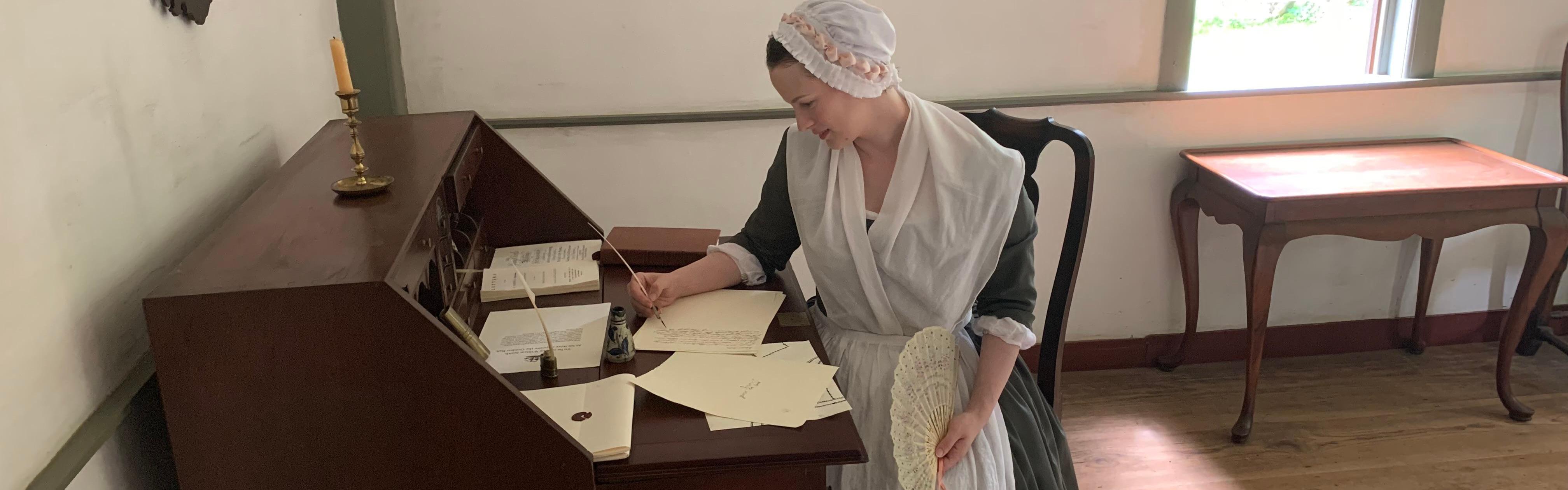 A woman in colonial clothing sits at a desk writing