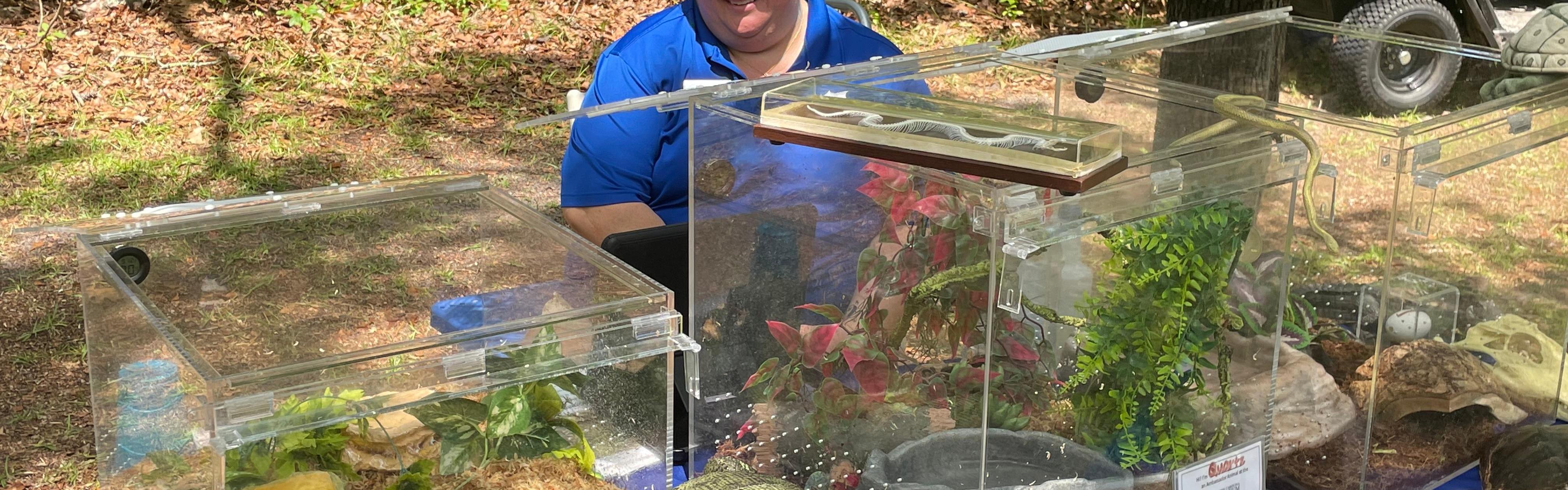 Woman in a blue polo shirt sits at a table with a variety of amphibians and reptiles in glass cages