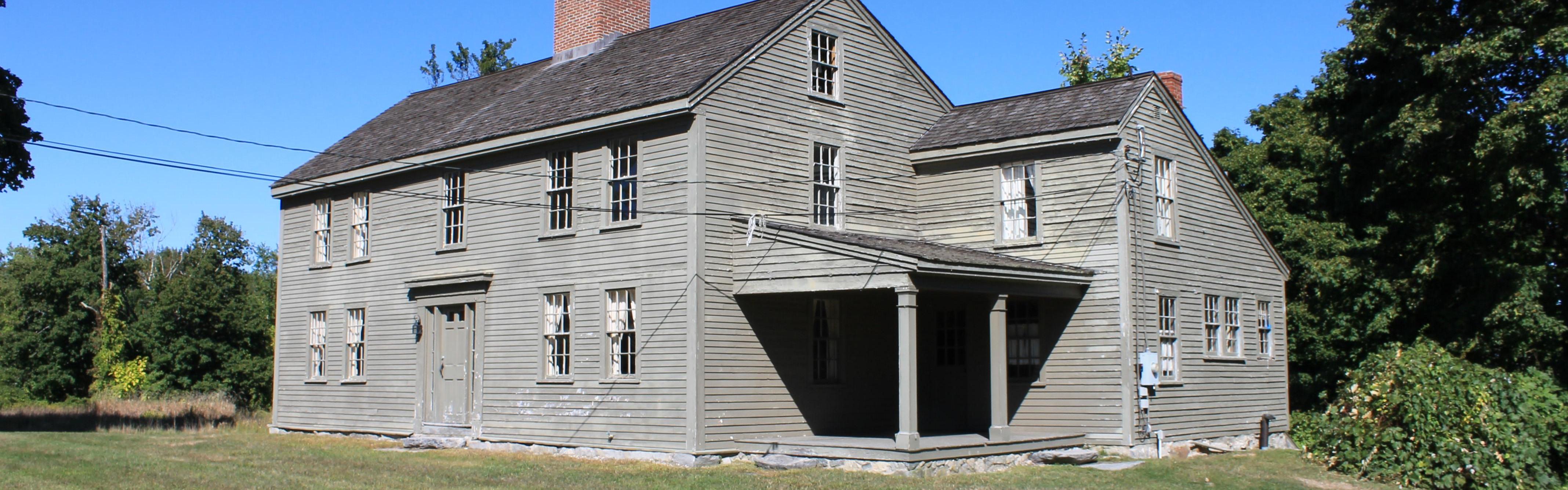 A wooden 18th century house sits in a field of green grass