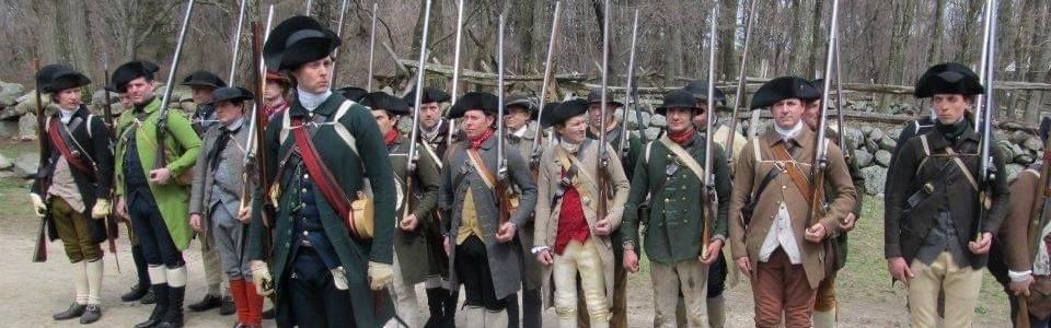 Several Colonial Revolutionary War militiamen stand in a line with muskets.