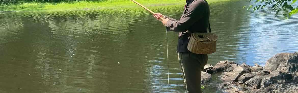 An 18th century man stands at a river fishing. He has a wicker basket on his hip and no shoes on.