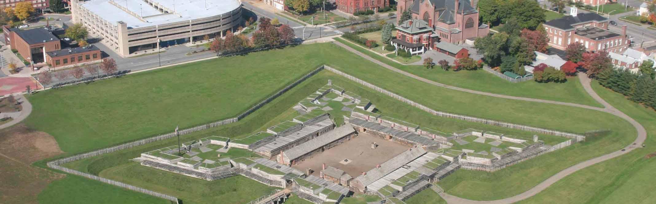 A view of a star-shaped wooden fort in a field of verdant green, from above. A city surrounds it.