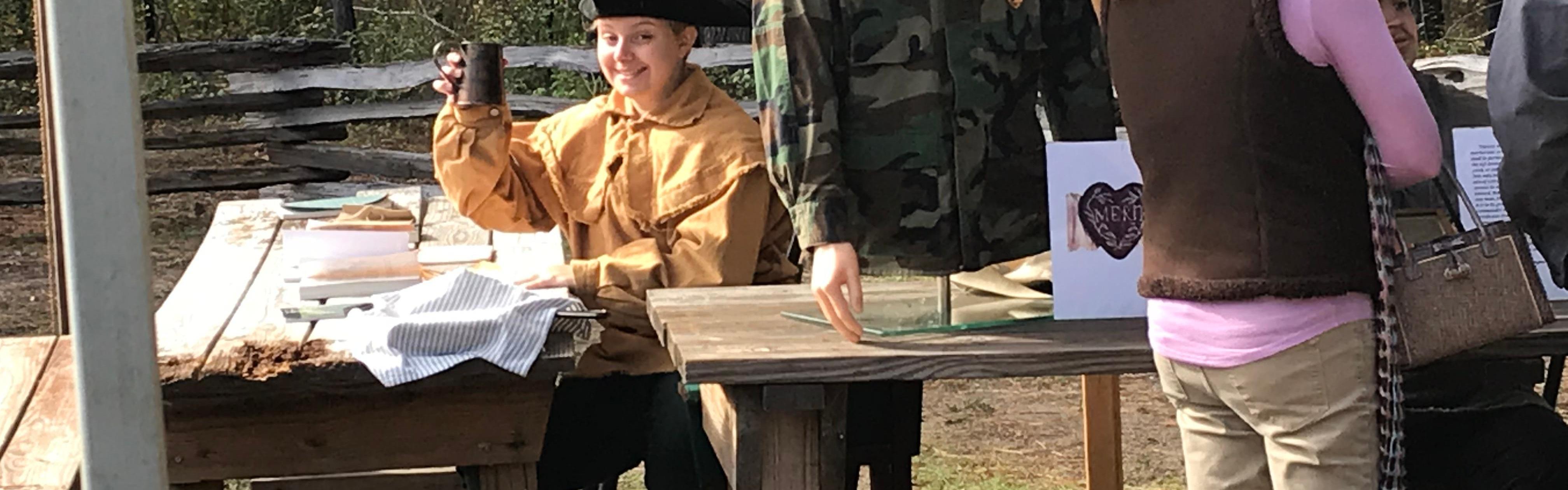 Person wearing colonial clothing sitting at a wooden table with a mannequin on the table.