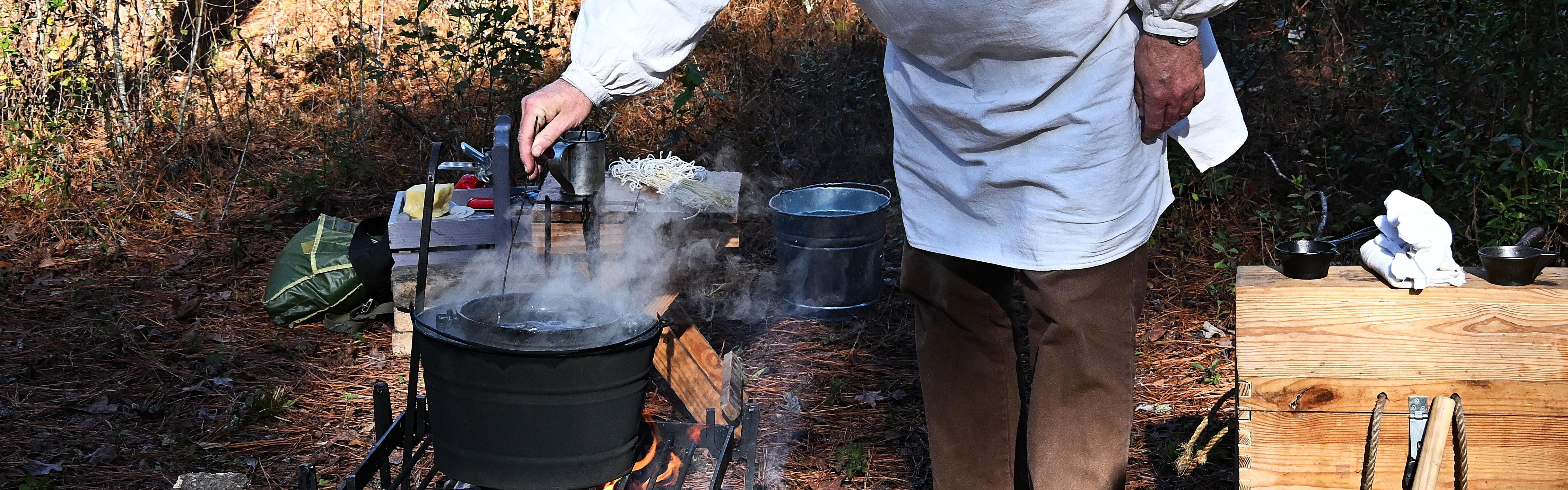 Man in a white smock bending over a campfire dipping a candle