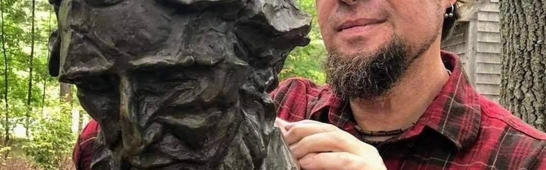 A man in a plaid shirt leans on a bronze monument of Henry David Thoreau