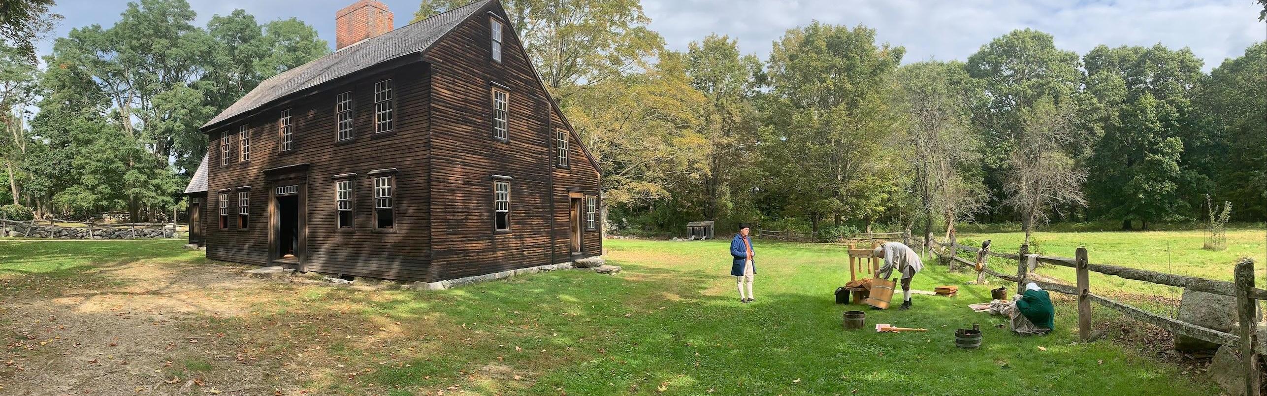 A historic wooden house in a field with people dressed in colonial clothing outside, and a cider pre