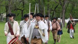 Men in colonial clothing marching behind a drummer.