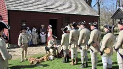 Men in grey Continental soldier uniforms stand in line near a large wooden building.