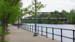 Along a sidewalk, you can see a metal railing and just beyond that, a river and a metal bridge.