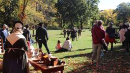 People in colonial clothing mingle with modern visitors in a field with trees in autumn color
