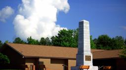 US Monument stands in front of visitor center. White cloud in deep blue sky is above them.