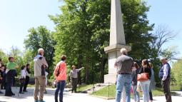 A park ranger stands in front of a towering stone monument pointing as visitors stand around.