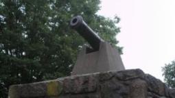 A black cannon sits atop a small stone structure with a glass window, surrounded by green trees under an overcast sky.