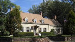 A tan colonial‑style house with dormer windows, brick chimneys, and a columned porch stands behind trimmed hedges and a stone retaining wall, with an American flag flying beside it.