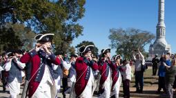 Fifes and drum wearing colonial uniforms with red jackets, march by the Yorktown Victory monument