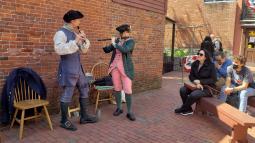 Two people wearing colonial garb and playing instruments while a few people watch