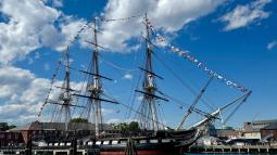 USS Constitution, a three-masted sailing ship, is decorated in flags on a partly-cloudy day.