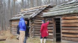 Two men in colonial living history clothing standing in front of log cabins.
