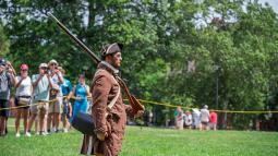 A ranger dressed in period clothing has a musket resting on his shoulder in front of an audience