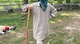 A colonial man with a cloth over his face stands holding a shovel