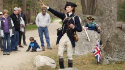 A Park Ranger in 18th Militia clothing stands in front of a crowd talking and pointing.