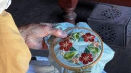 A volunteer in colonial dress practices cross stich. She is stitching a flower design.