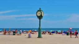 Green clock on boardwalk of ocean beach park site with people, umbrellas and blankets on sand