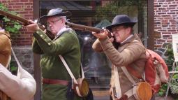 two men in colonial militia clothing pointing muskets at the camera