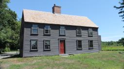 A wooden two story colonial house with dark paint sits in a field of green grass