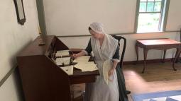 A woman in colonial clothing sits at a desk writing