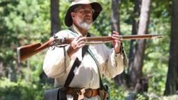 A living historian holds a weapon with trees behind him.