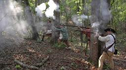 Living historians fire muskets with gray smoke coming from them. They are in the woods.