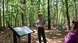 Man in green and gray NPS uniform standing next to an exhibit panel talking with visitors