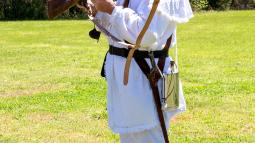 A man dressed in while colonial clothes with a black tri-corn hat discusses a musket.