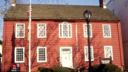A red historic house with white trim and twin brick chimneys stands behind a flagpole and walkway lined with bushes.