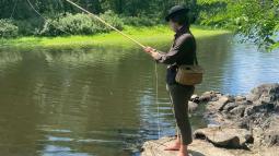 An 18th century man stands at a river fishing. He has a wicker basket on his hip and no shoes on.