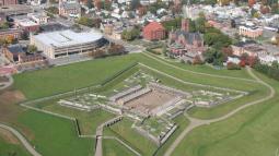 A view of a star-shaped wooden fort in a field of verdant green, from above. A city surrounds it.